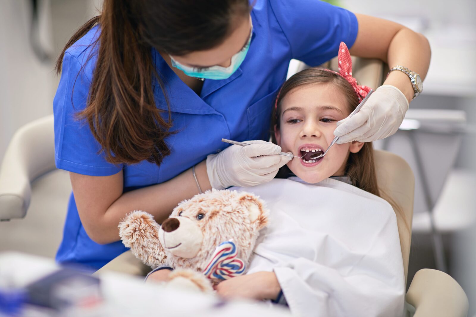 A children’s dentist in Montebello carefully examining a young girl’s teeth while she holds a stuffed teddy bear, seated comfortably in a modern dental chair.