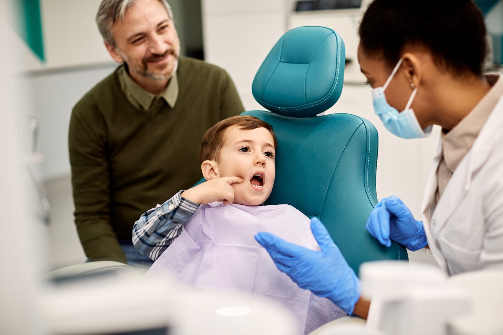 A general dentist in Montebello attending to a young child seated in a teal dental chair, with a guardian watching nearby in a modern dental office. The dentist wears a mask and gloves while engaging with the child, ensuring a comfortable and friendly environment.